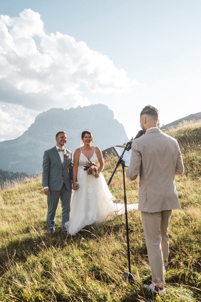 Freie Trauung auf einer Almwiese in den Dolomiten, Brautpaar lacht während Alex spricht
