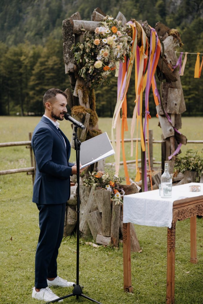 Freier Redner Alex am Mikrofon vor einem Holz-Traubogen mit bunten Bändern und Wiesenblumen