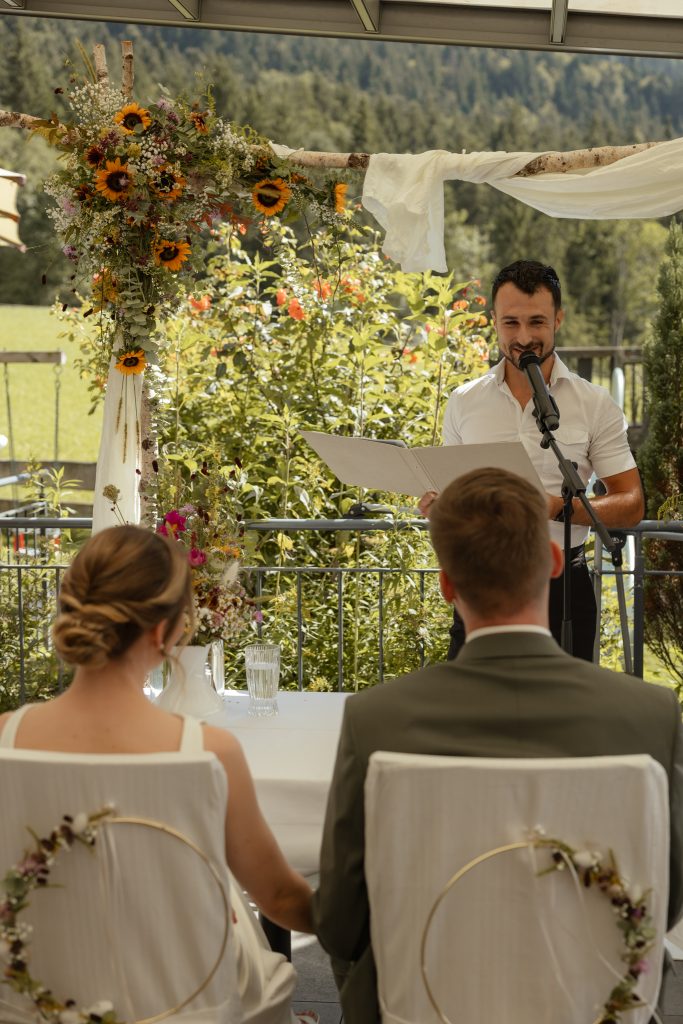 Freie Trauung auf einer Terrasse mit Sonnenblumen-Traubogen und Bergwald im Hintergrund